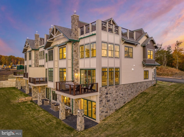 back of house featuring a balcony, stone siding, central AC, and a lawn