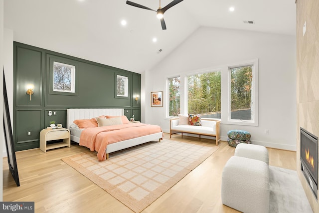 bedroom featuring high vaulted ceiling, light wood-type flooring, a tiled fireplace, and ceiling fan