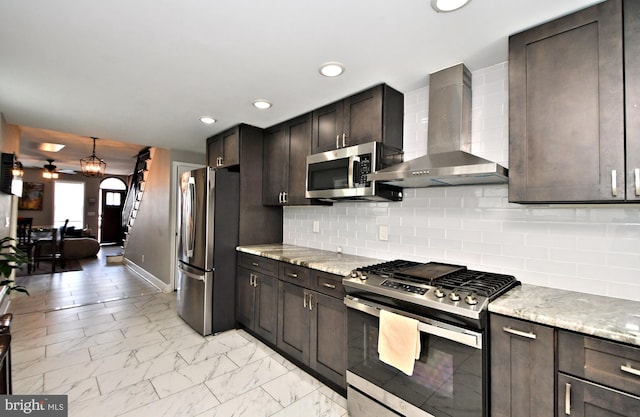 kitchen featuring dark brown cabinetry, light stone countertops, stainless steel appliances, wall chimney range hood, and decorative backsplash