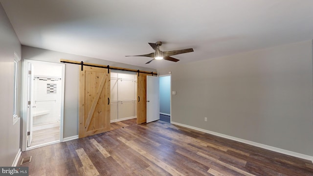 unfurnished bedroom featuring ensuite bathroom, ceiling fan, a barn door, dark wood-type flooring, and a closet