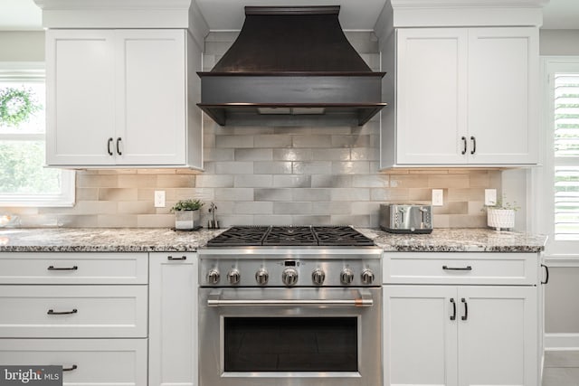 kitchen with custom exhaust hood, high end stainless steel range, tasteful backsplash, light stone counters, and white cabinetry