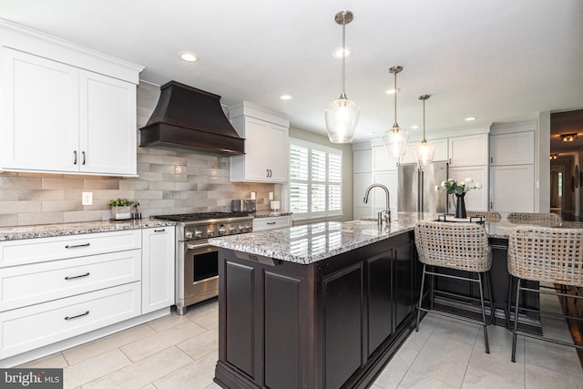 kitchen featuring custom range hood, premium appliances, a kitchen island with sink, white cabinetry, and hanging light fixtures