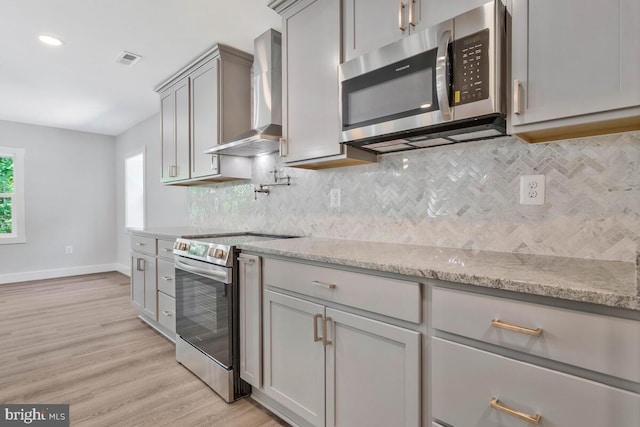 kitchen featuring gray cabinetry, light stone countertops, wall chimney exhaust hood, stainless steel appliances, and light hardwood / wood-style flooring