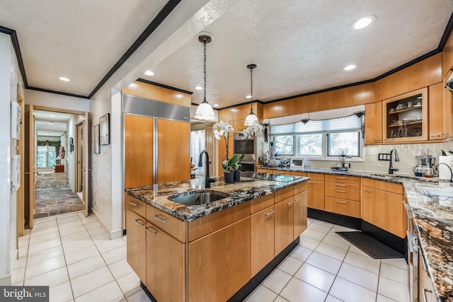 kitchen featuring crown molding, sink, a center island with sink, dark stone countertops, and hanging light fixtures