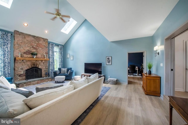 living area featuring light wood-type flooring, high vaulted ceiling, a stone fireplace, a skylight, and baseboards