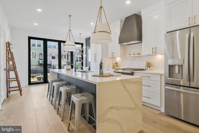 kitchen featuring wall chimney range hood, hanging light fixtures, a center island with sink, white cabinets, and appliances with stainless steel finishes