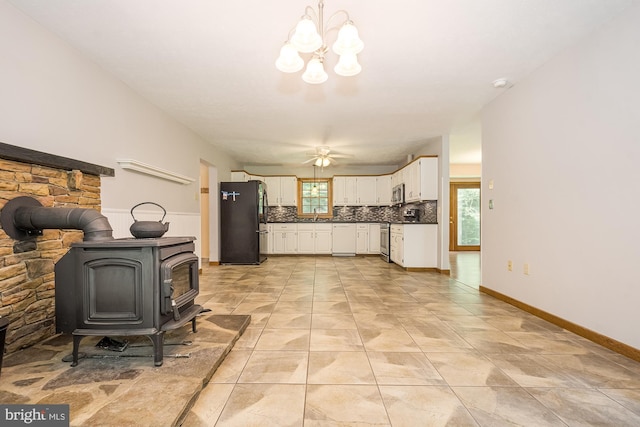 kitchen with a wood stove, stainless steel appliances, decorative backsplash, white cabinets, and ceiling fan with notable chandelier