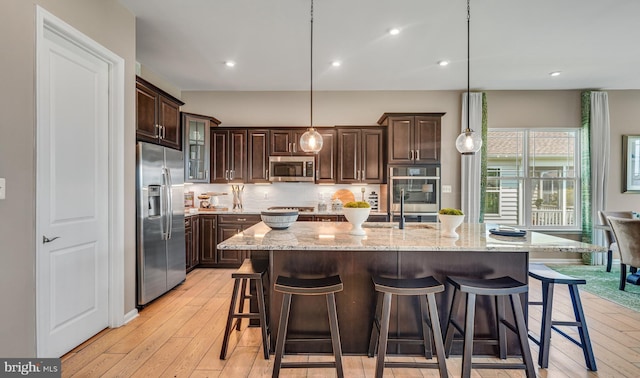 kitchen featuring pendant lighting, stainless steel appliances, light stone counters, dark brown cabinetry, and an island with sink