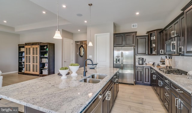 kitchen with appliances with stainless steel finishes, light wood-type flooring, a kitchen island with sink, sink, and hanging light fixtures