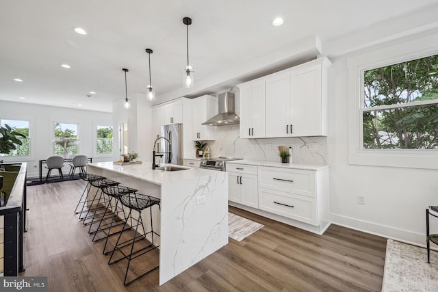 kitchen with wall chimney range hood, white cabinets, dark wood-type flooring, stainless steel appliances, and a center island with sink