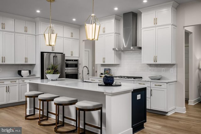 kitchen featuring white cabinetry, wall chimney range hood, stainless steel appliances, hanging light fixtures, and a center island with sink