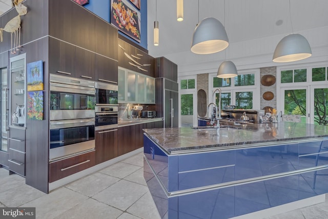 kitchen with stainless steel double oven, dark stone countertops, decorative light fixtures, a center island with sink, and dark brown cabinets