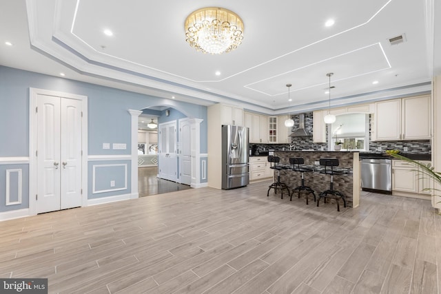 kitchen featuring hanging light fixtures, wall chimney exhaust hood, light wood-type flooring, appliances with stainless steel finishes, and a tray ceiling