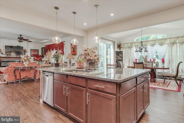 kitchen with hardwood / wood-style floors, stainless steel dishwasher, ceiling fan, an island with sink, and light stone counters