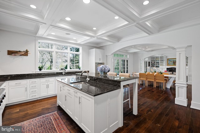 kitchen with ornate columns, sink, dark stone countertops, a center island with sink, and white cabinets