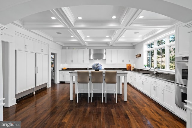 kitchen featuring coffered ceiling, wall chimney range hood, double oven, a kitchen island, and white cabinetry