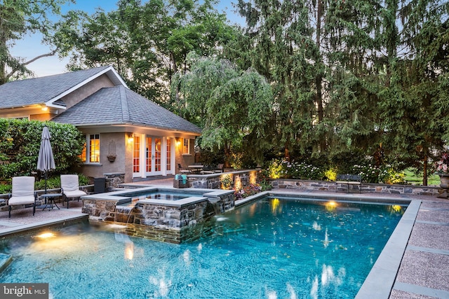 pool at dusk with an in ground hot tub, pool water feature, an outdoor structure, and a patio