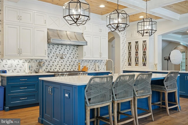 kitchen with blue cabinetry, an island with sink, beam ceiling, and custom exhaust hood