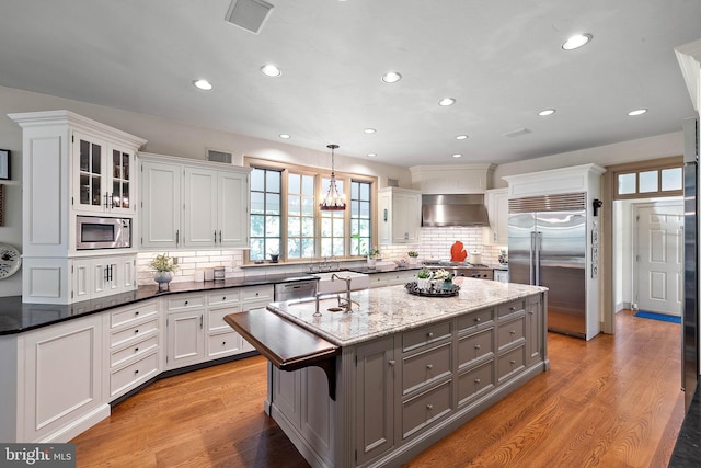 kitchen featuring built in appliances, wall chimney exhaust hood, dark stone countertops, decorative light fixtures, and white cabinetry