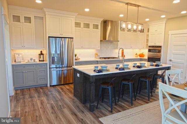 kitchen featuring stainless steel appliances, wall chimney range hood, a center island with sink, dark hardwood / wood-style floors, and white cabinetry