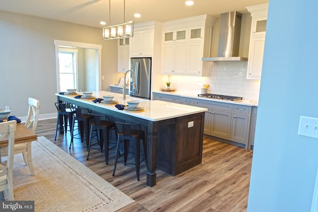 kitchen with white cabinets, wall chimney range hood, an island with sink, appliances with stainless steel finishes, and decorative light fixtures