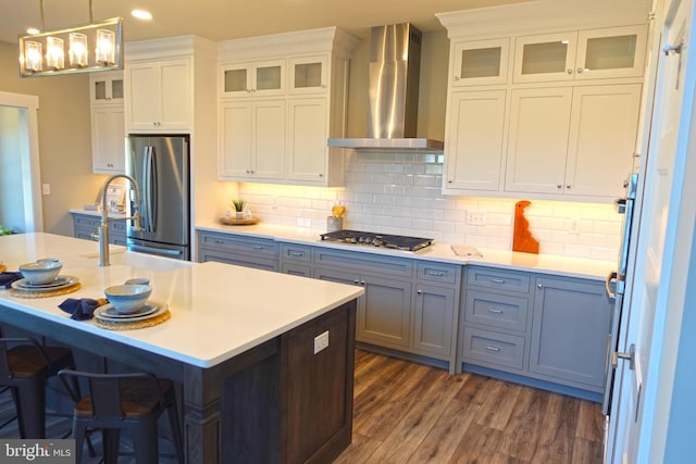 kitchen featuring wall chimney exhaust hood, tasteful backsplash, an island with sink, white cabinets, and appliances with stainless steel finishes