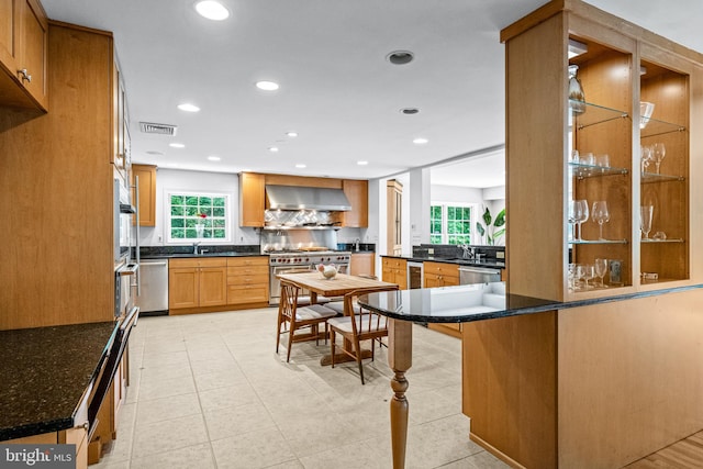 kitchen with light tile patterned floors, kitchen peninsula, dark stone counters, wall chimney range hood, and appliances with stainless steel finishes