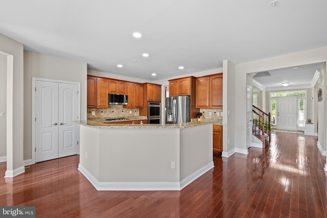 kitchen with light stone counters, appliances with stainless steel finishes, backsplash, brown cabinetry, and dark wood finished floors