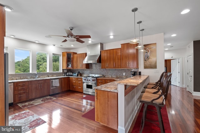 kitchen with kitchen peninsula, a kitchen breakfast bar, wall chimney exhaust hood, stainless steel appliances, and decorative light fixtures