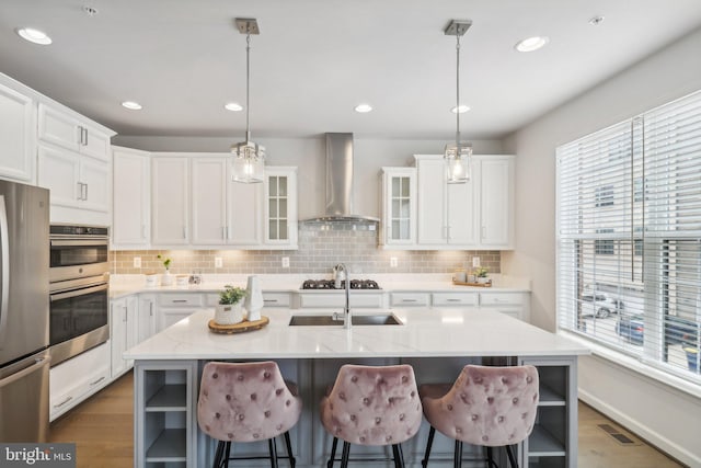 kitchen with pendant lighting, white cabinets, wall chimney exhaust hood, an island with sink, and appliances with stainless steel finishes