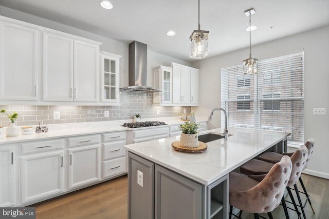 kitchen featuring stainless steel gas stovetop, a kitchen island with sink, sink, wall chimney exhaust hood, and white cabinetry