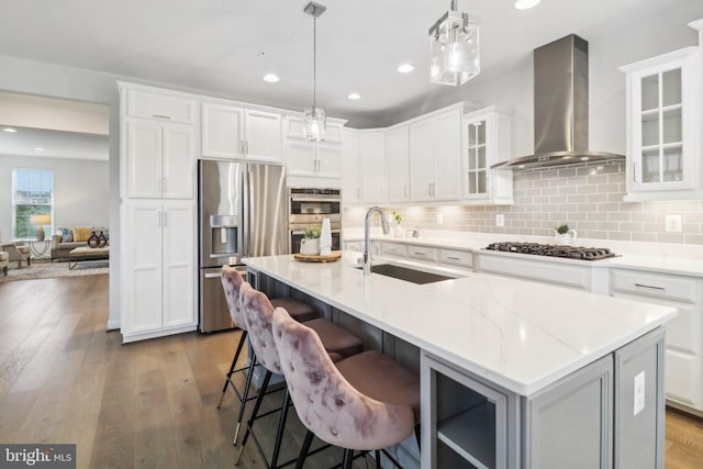 kitchen featuring wall chimney exhaust hood, an island with sink, decorative light fixtures, white cabinetry, and stainless steel appliances