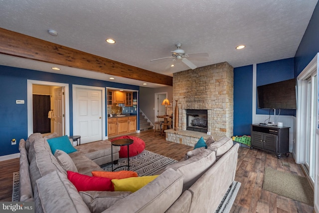 living room featuring dark wood-type flooring, a stone fireplace, ceiling fan, a textured ceiling, and beamed ceiling