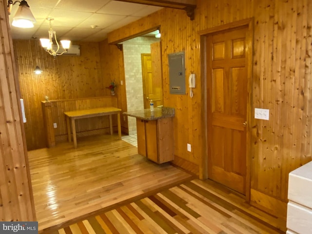 corridor with light wood-type flooring, a drop ceiling, a notable chandelier, electric panel, and wood walls