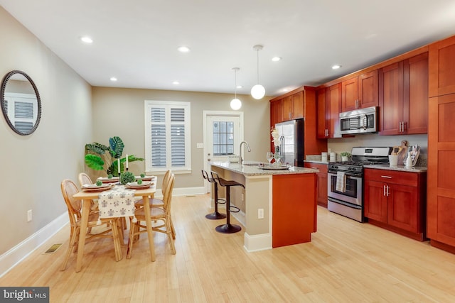 kitchen featuring light stone countertops, an island with sink, appliances with stainless steel finishes, decorative light fixtures, and light hardwood / wood-style floors