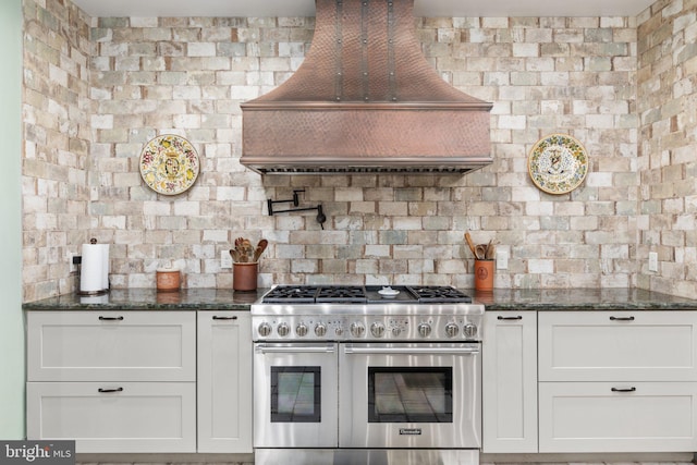 kitchen with custom exhaust hood, dark stone countertops, white cabinets, and double oven range