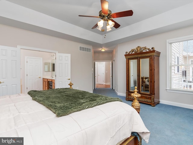 carpeted bedroom featuring a tray ceiling, ensuite bathroom, and ceiling fan