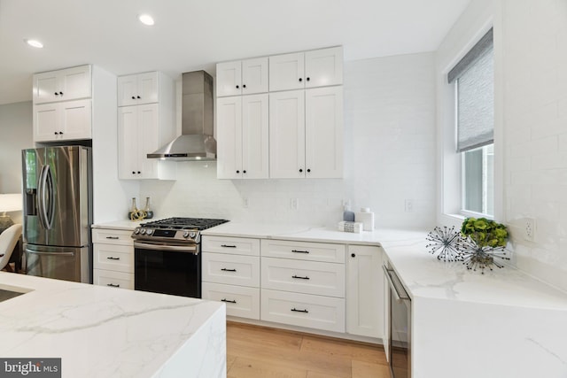 kitchen with light hardwood / wood-style floors, stainless steel appliances, white cabinets, wall chimney exhaust hood, and light stone counters