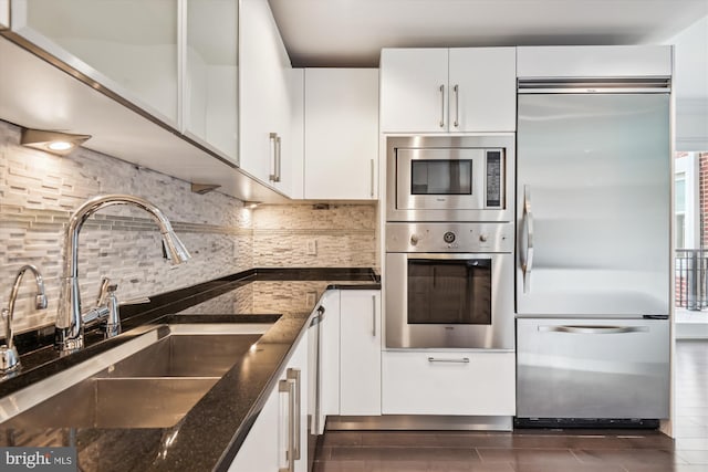 kitchen featuring sink, backsplash, built in appliances, dark stone counters, and white cabinets