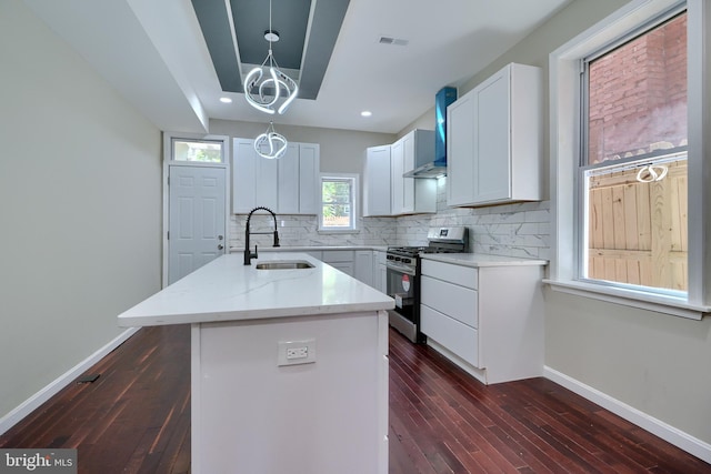 kitchen with stainless steel gas range oven, wall chimney range hood, sink, white cabinets, and an island with sink