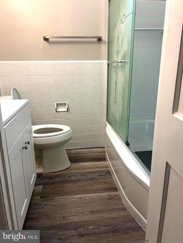 full bathroom featuring vanity, bath / shower combo with glass door, hardwood / wood-style flooring, toilet, and tile walls