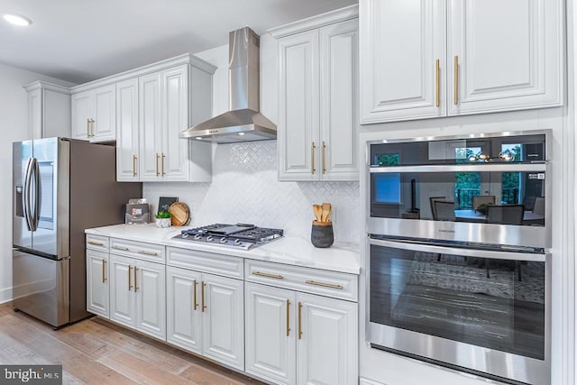 kitchen with wall chimney exhaust hood, stainless steel appliances, decorative backsplash, light wood-type flooring, and white cabinetry