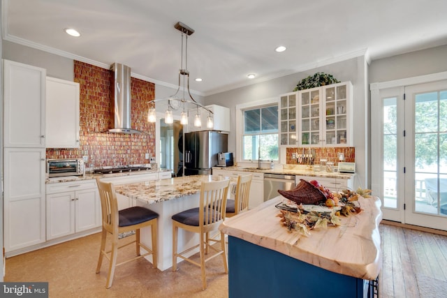 kitchen featuring stainless steel appliances, wall chimney range hood, decorative light fixtures, white cabinets, and a center island