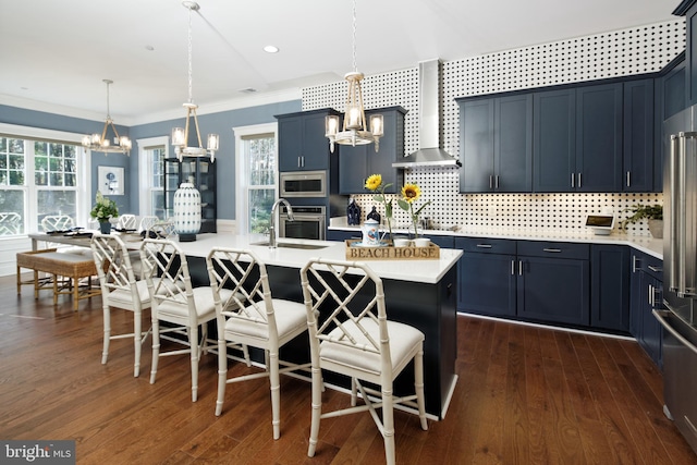 kitchen with hanging light fixtures, wall chimney range hood, decorative backsplash, a center island with sink, and appliances with stainless steel finishes