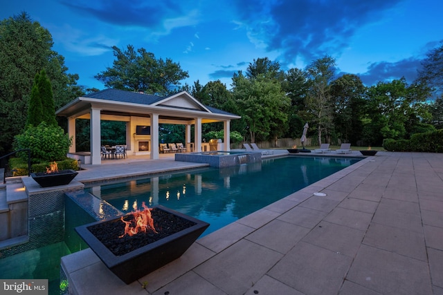 pool at dusk featuring an in ground hot tub, exterior fireplace, a gazebo, an outdoor fire pit, and a patio