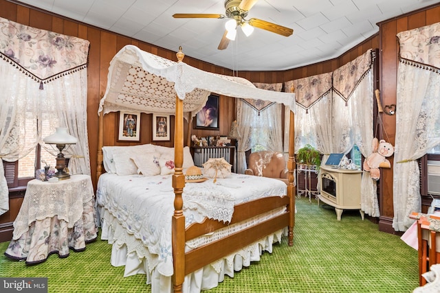 bedroom featuring carpet flooring, a wood stove, ceiling fan, and wooden walls