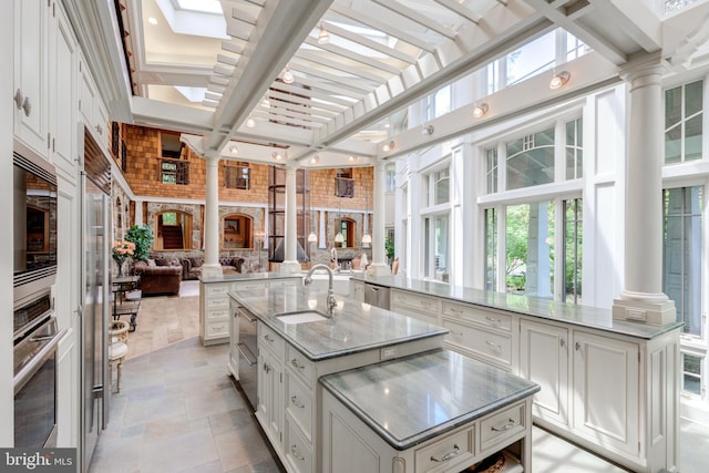 kitchen with light stone countertops, ornate columns, stainless steel appliances, sink, and a large island with sink