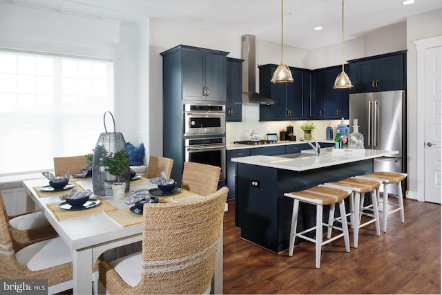 kitchen featuring hanging light fixtures, wall chimney exhaust hood, an island with sink, appliances with stainless steel finishes, and a kitchen bar