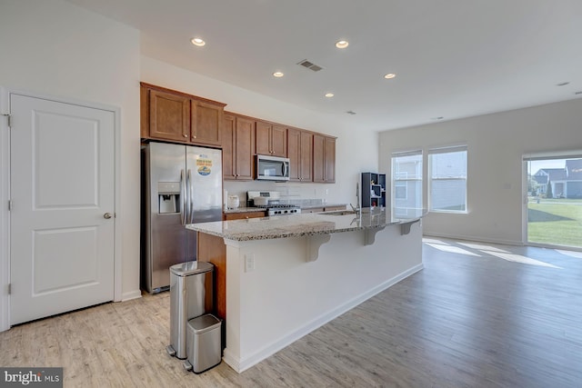 kitchen with a center island with sink, appliances with stainless steel finishes, light stone countertops, and light wood-type flooring