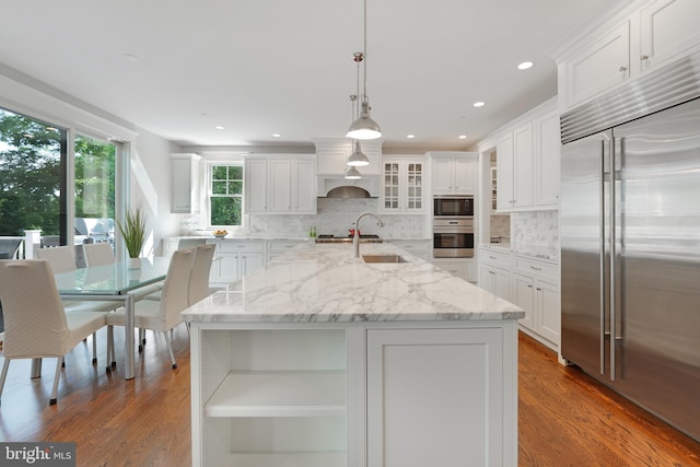 kitchen featuring white cabinets, built in appliances, hanging light fixtures, and an island with sink